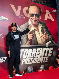 Un hombre señala un cartel con un personaje interpretado por él mismo.