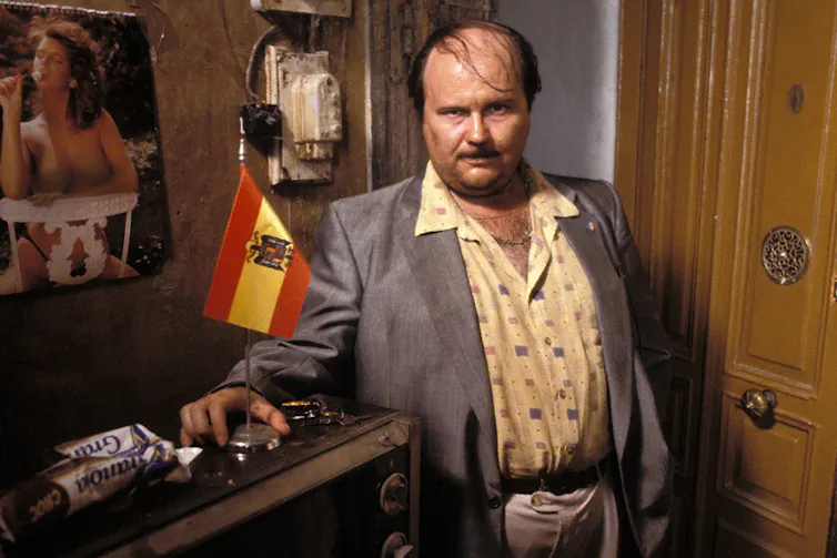 Un hombre gordo posa delante de una mesa con la bandera de España y un póster de una mujer desnuda en la pared.