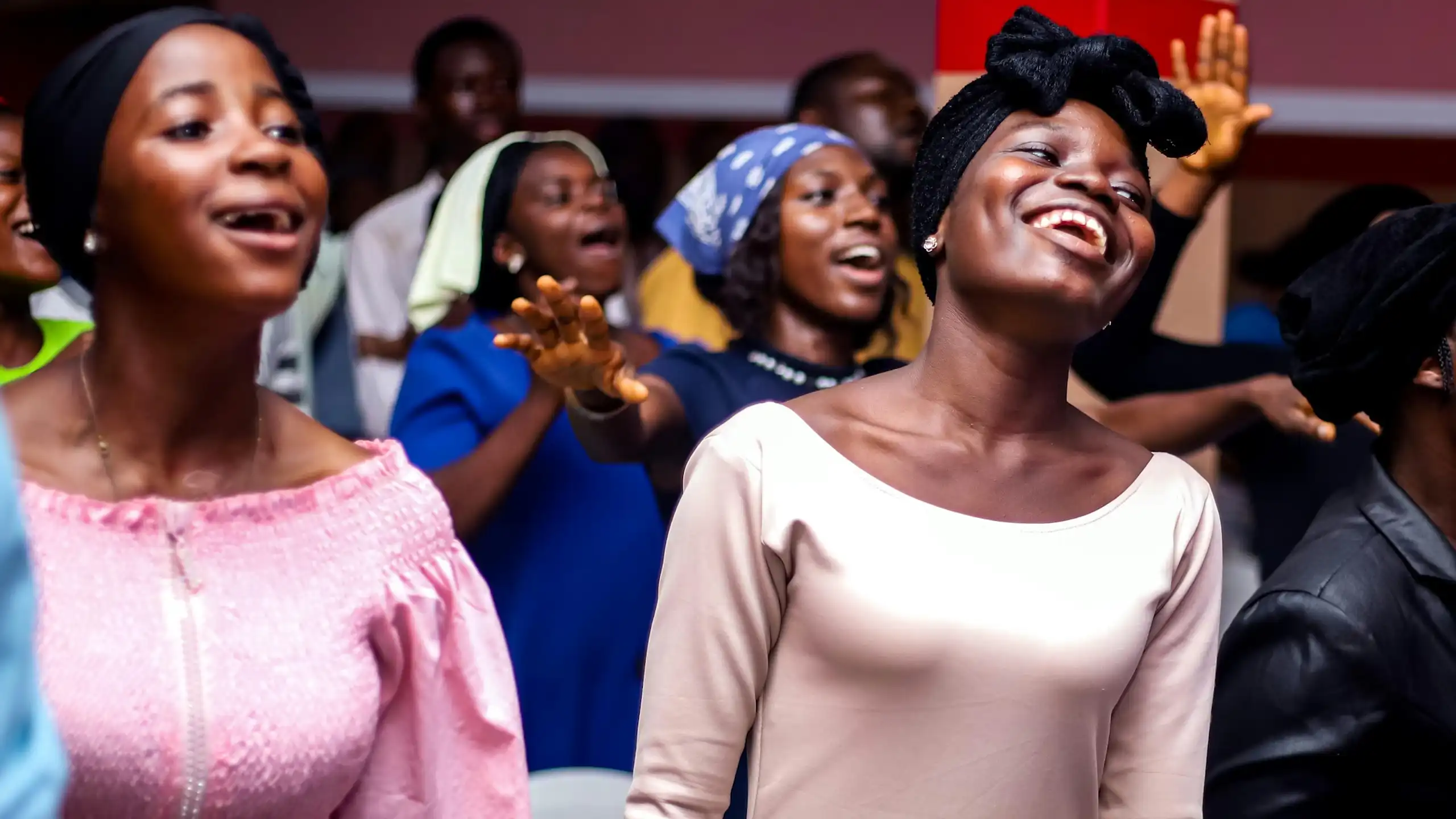 Two young African women smile as they sing, surrounded by other people in a gathering, some with raised arms.