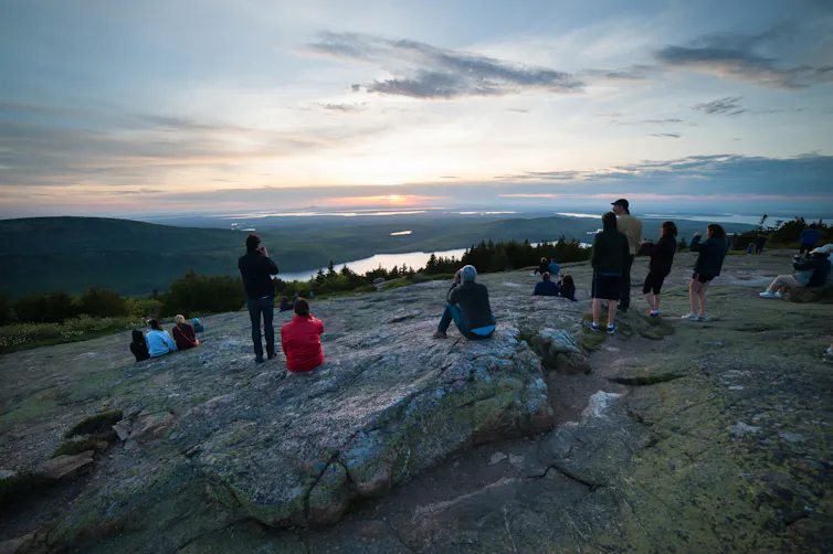 Este verano no estarás solo en los parques nacionales: disfruta de la compañía 1 Un grupo de personas se sienta y se para en la cima de una montaña y observa cómo sale el sol sobre el océano, el lago y algunas colinas más bajas.