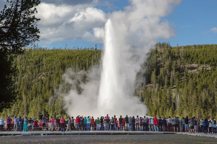 A large group of people stand in a row watching a large spout of water with a wooded hillside in the background.