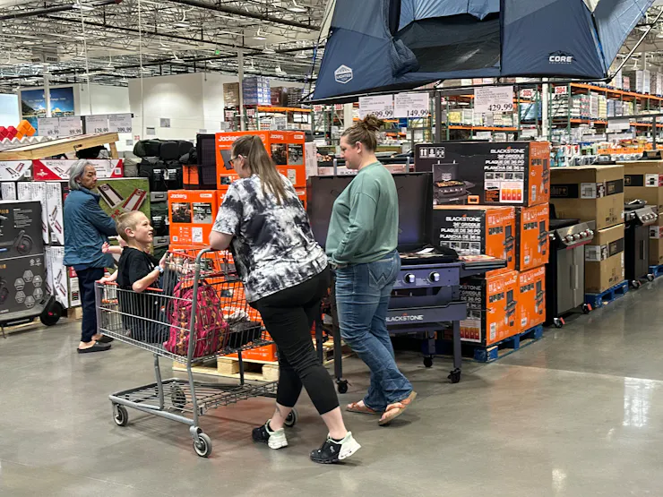 Several women shoppers and a child in a shopping card are looking at outdoor cooking equipment on display in a Costco warehouse.