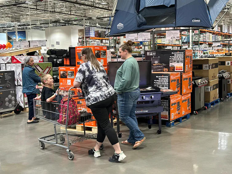 Several women shoppers and a child in a shopping card are looking at outdoor cooking equipment on display in a Costco warehouse.