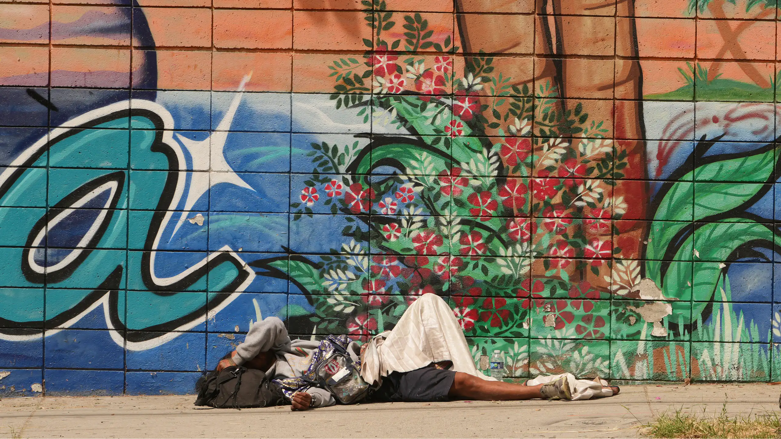 A homeless man lies on the sidewalk, next to artistic graffiti.