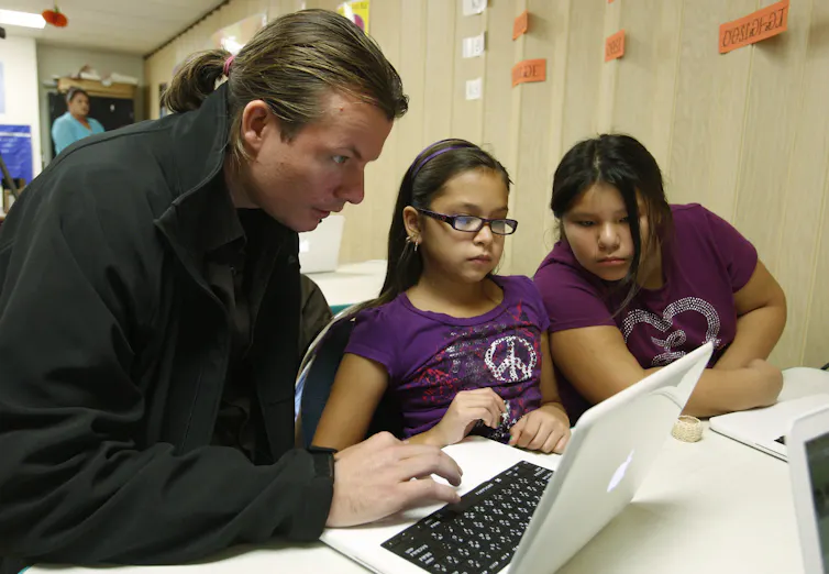 A man is using a laptop to teach two young girls seated next to him.