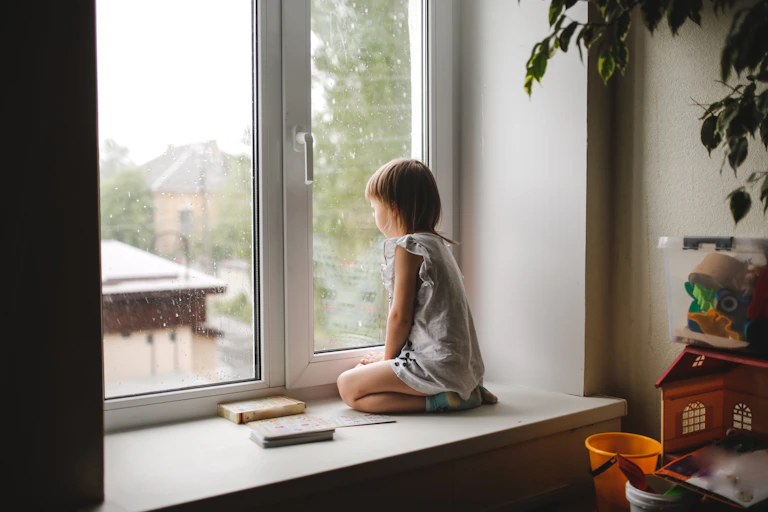 A young child sits on a ledge, looking out of a big window.
