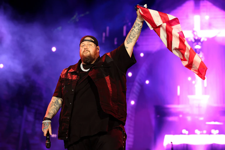 A heavily-tattooed singer in black clothes waves the US flag onstage at a concert.