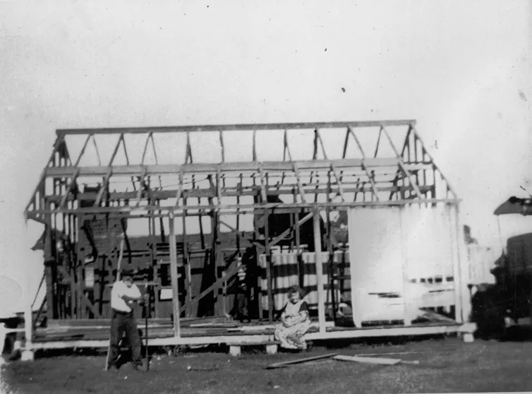 A black and white image of a timber frame house under construction with a man and woman standing and sitting in front.