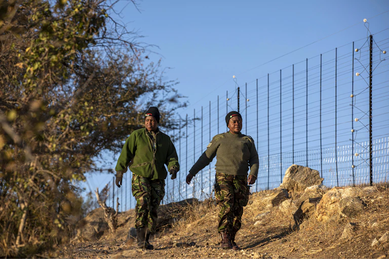 Two women in green uniforms walk next to a tall fence