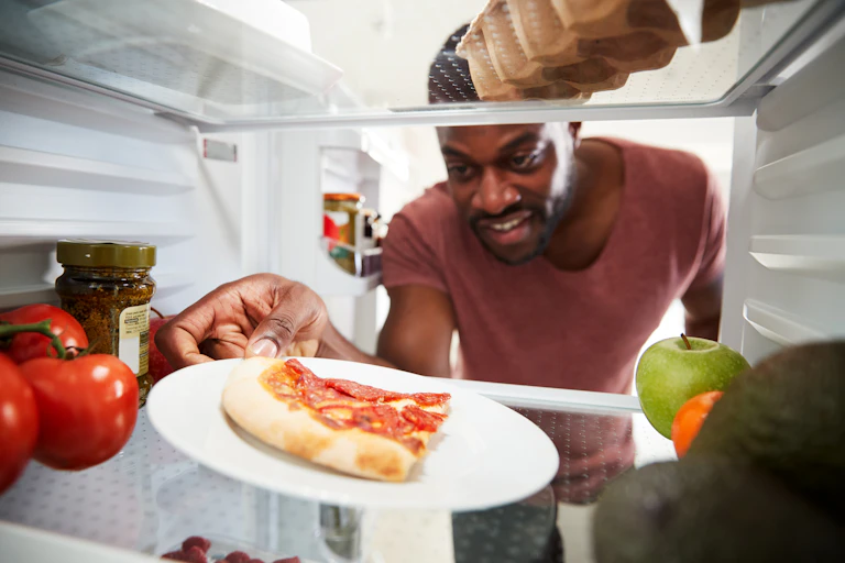 A man reaches into a fridge and grabs a slice of leftover, cold pizza that's sitting on a white plate.