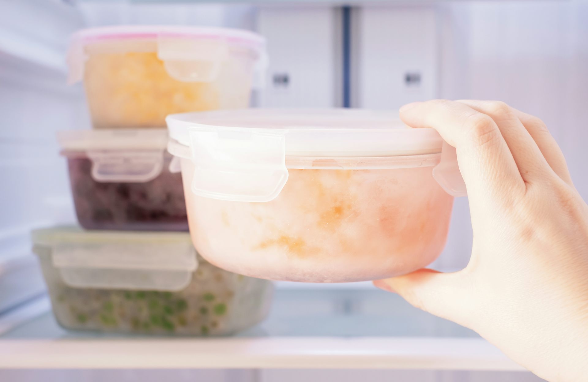 A person places a plastic tupperware container full of leftovers in the fridge next to three other containers of leftovers.