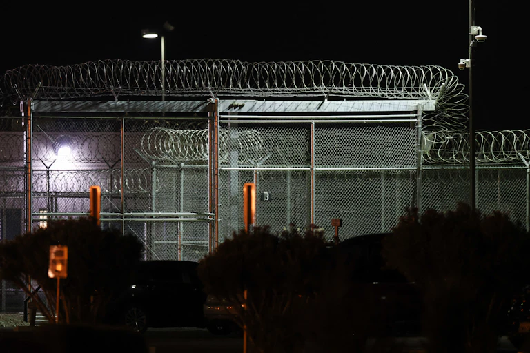 A large building surrounded by barbed wire is lit up by strong lights in the night.