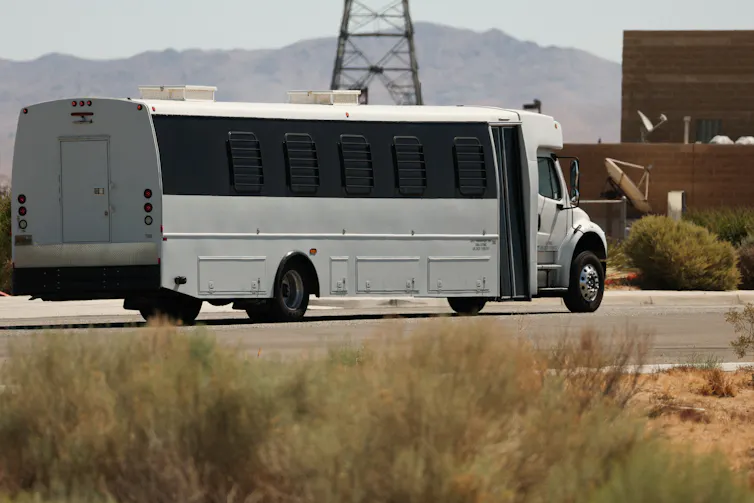 A white bus with blacked-out windows drives towards a large building.