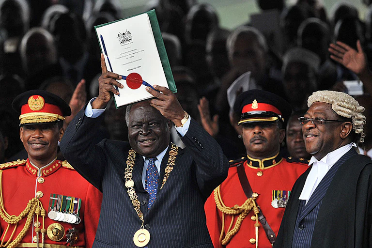 Man dressed in a suit with a gold mace around his neck holding up a document while other people around him smile