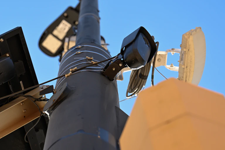 A street light pole with a solar panel, pedestrian signals and a small, black camera affixed to it.