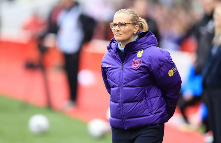 Sarina Wiegman stands on a football pitch in a purple puffer jacket.