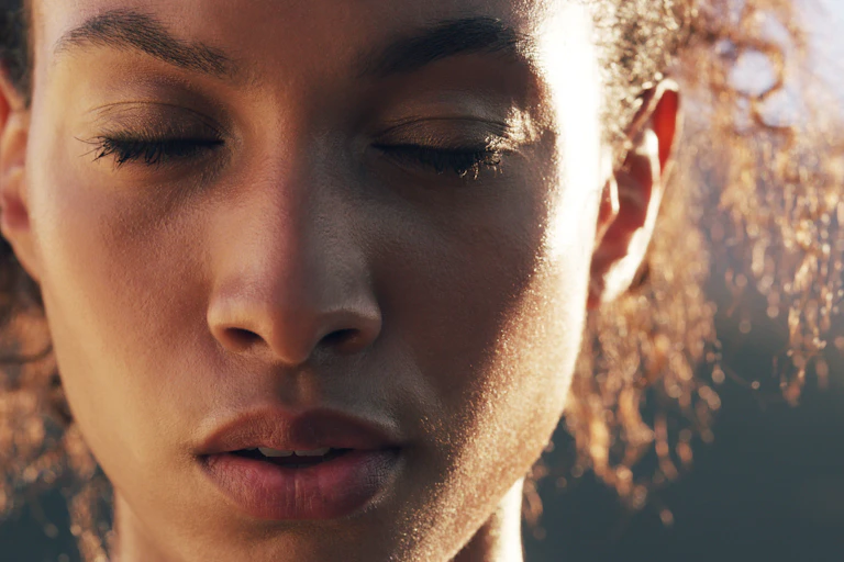 Close-up shot of a young woman closing her eyes, looking mindful, in an outdoor setting.