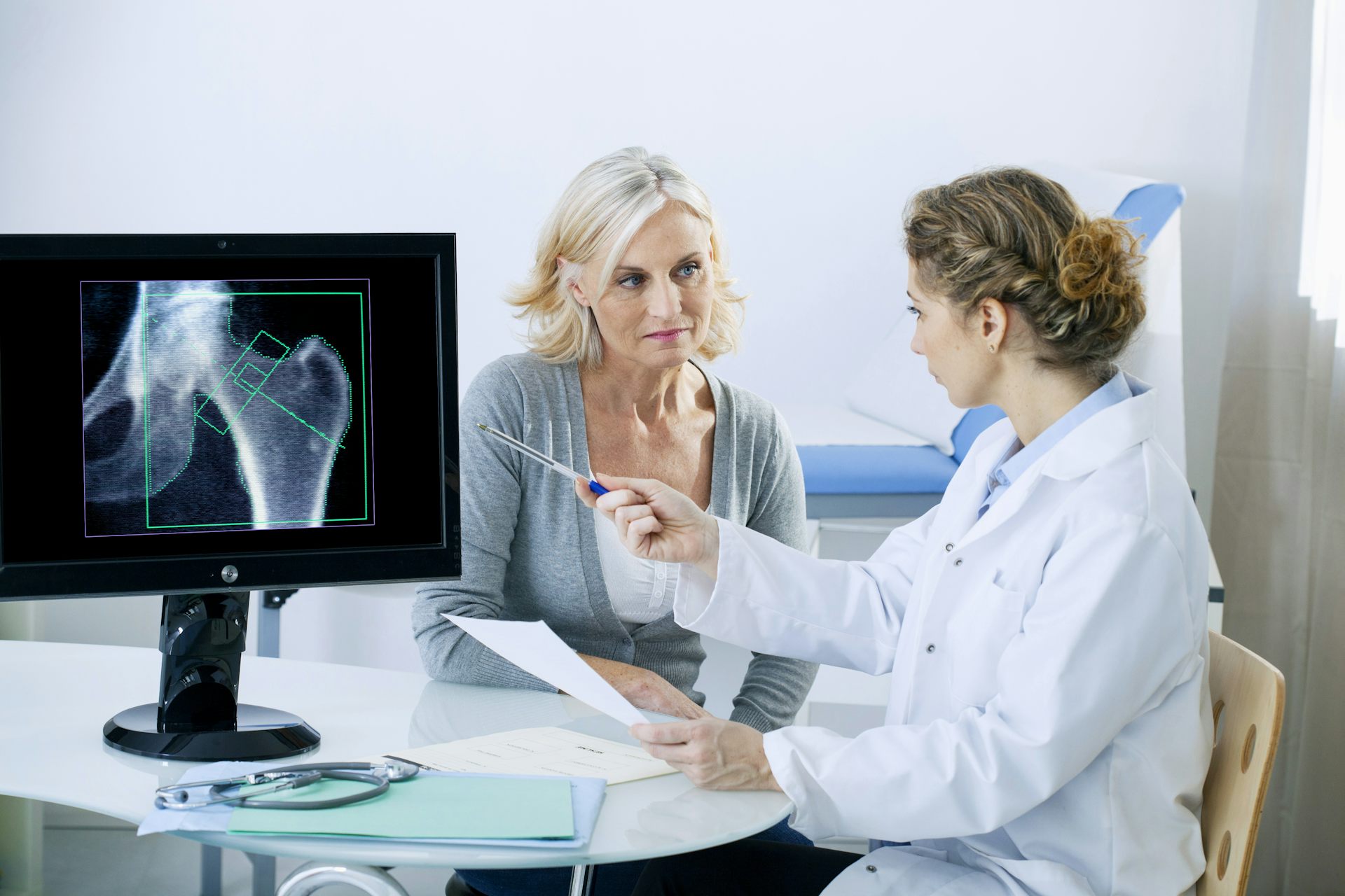An older woman listens to her doctor, while the female doctor explains the bone scan she has just has by pointing at the graphic on the computer screen.