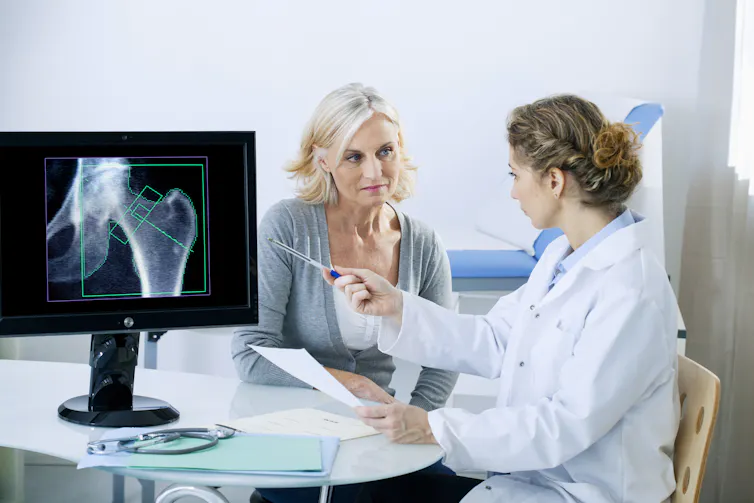 An older woman listens to her doctor, while the female doctor explains the bone scan she has just has by pointing at the graphic on the computer screen.
