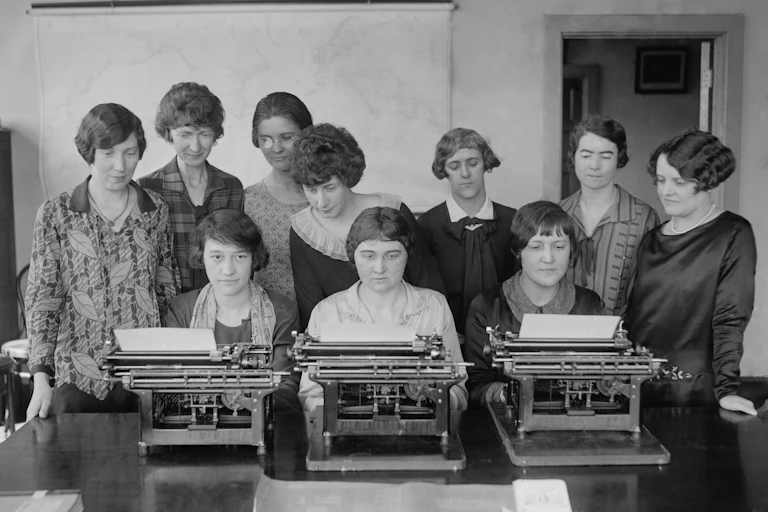 Un grupo de mujeres escriben mientras otras, de pie detrás de ella, las vigilan.