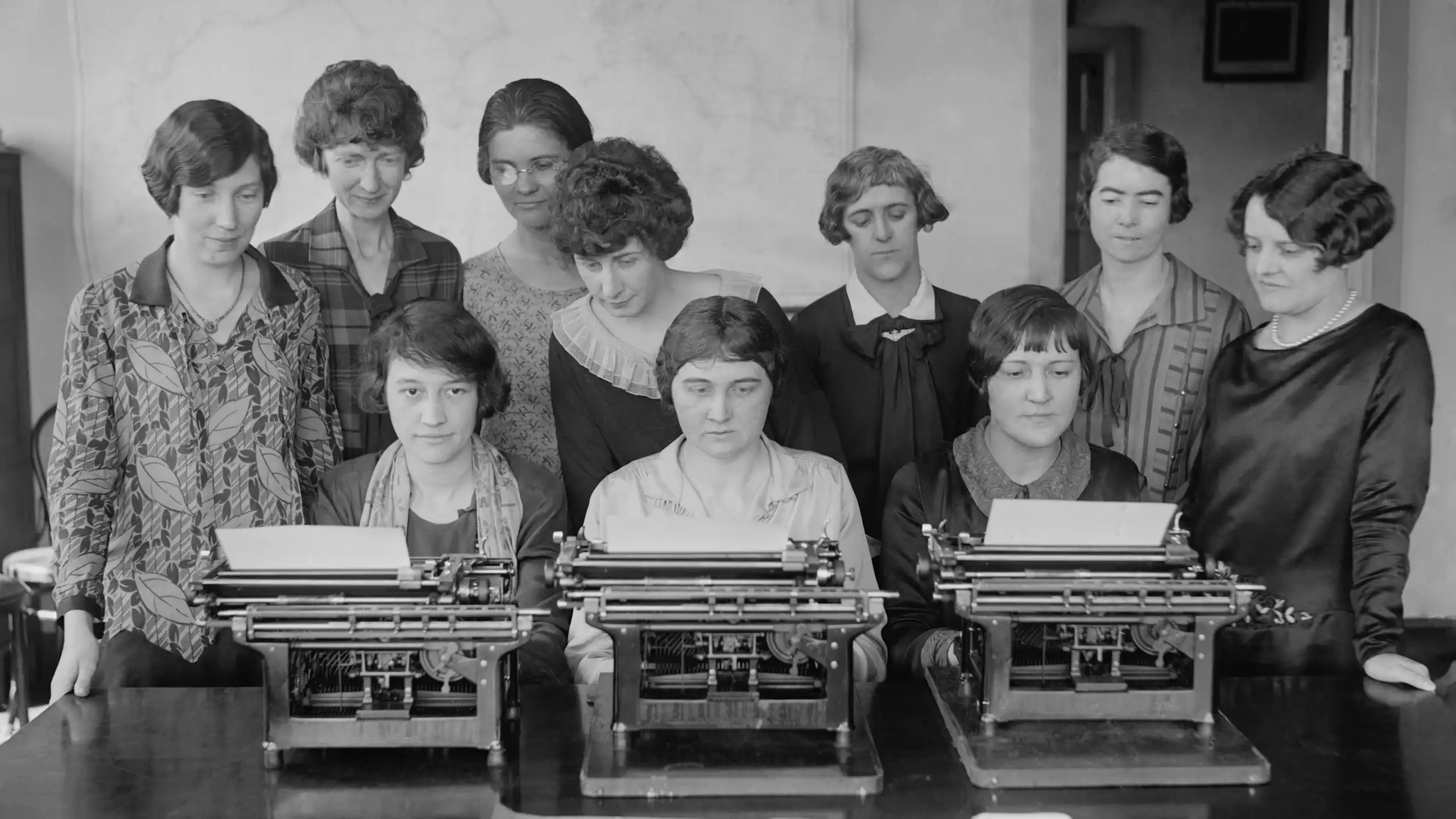 Un grupo de mujeres escriben mientras otras, de pie detrás de ella, las vigilan.