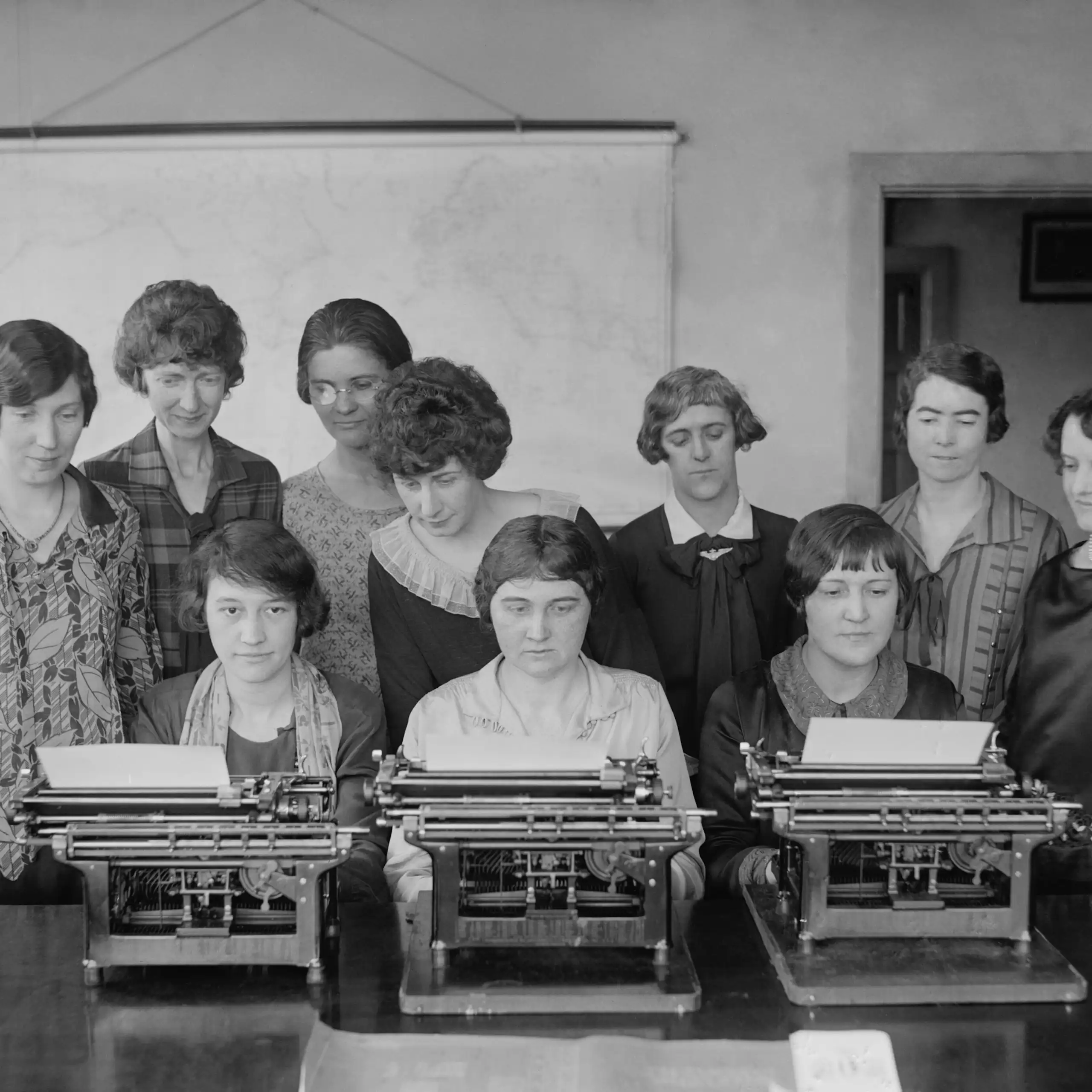 Un grupo de mujeres escriben mientras otras, de pie detrás de ella, las vigilan.