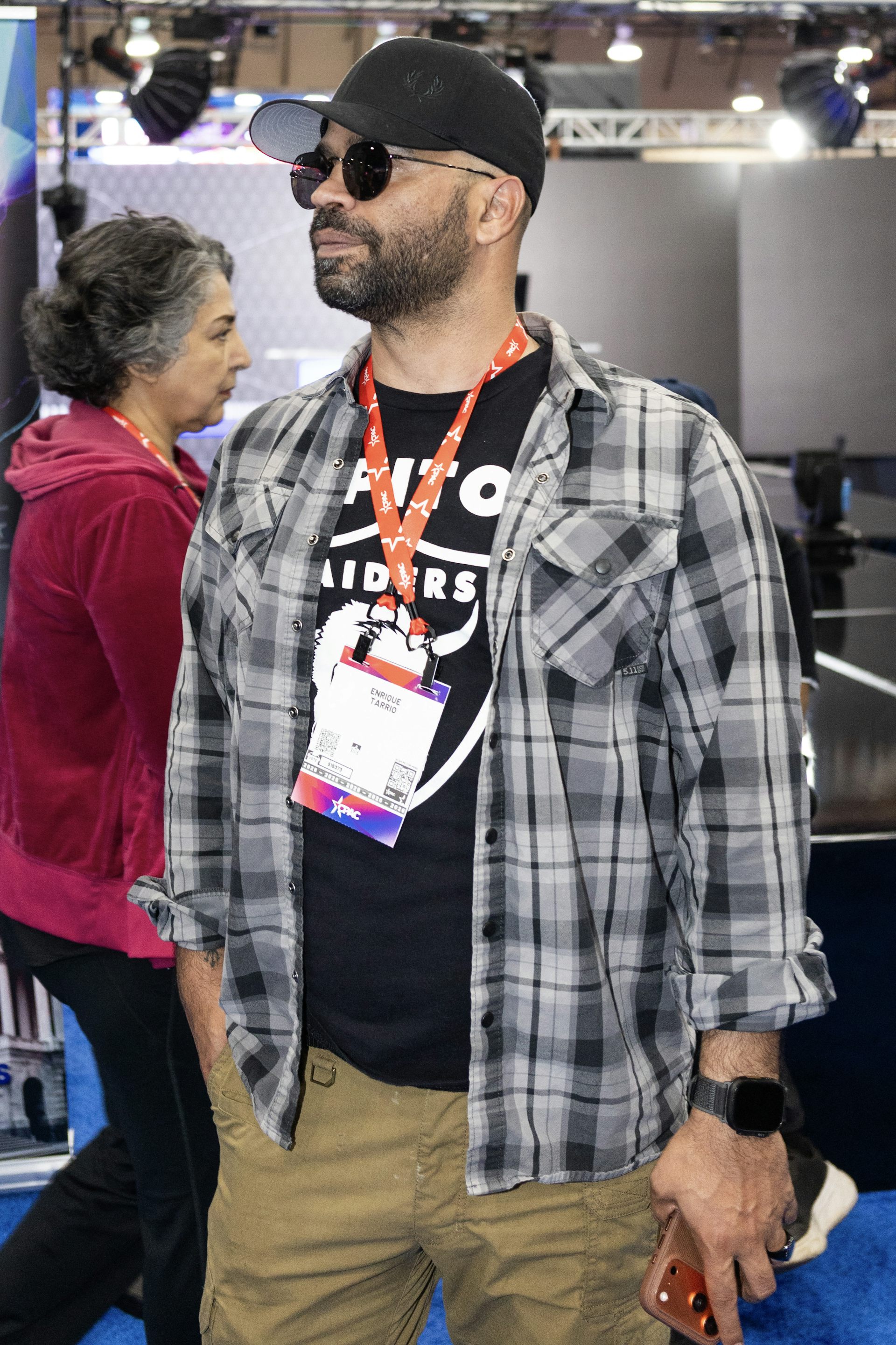 A man casually dressed in a t-shirt, plaid shirt over that, a black baseball cap and sunglasses stands in a convention center room.