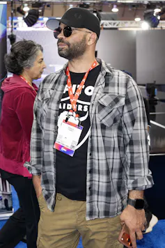 A man casually dressed in a t-shirt, plaid shirt over that, a black baseball cap and sunglasses stands in a convention center room.