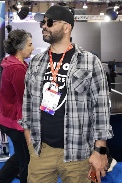 A man casually dressed in a t-shirt, plaid shirt over that, a black baseball cap and sunglasses stands in a convention center room.