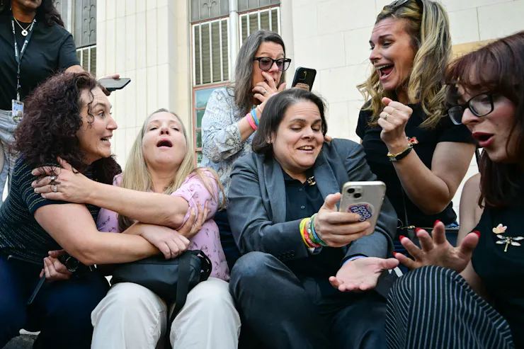 six woman seated outside a building have facial expressions indicating surprise and happiness