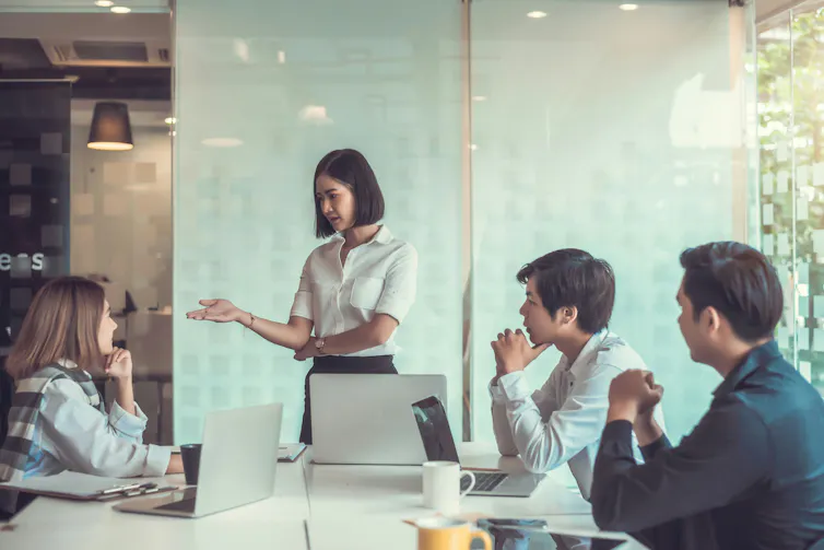 A woman, who is standing, speaks to a group of people sitting at a conference table