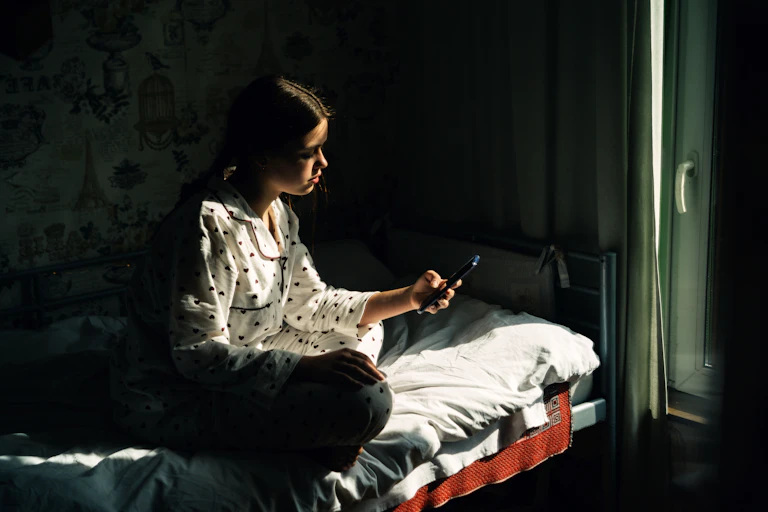 Uma adolescente vestindo pijama olha para o celular enquanto está sentada na cama em um quarto escuro