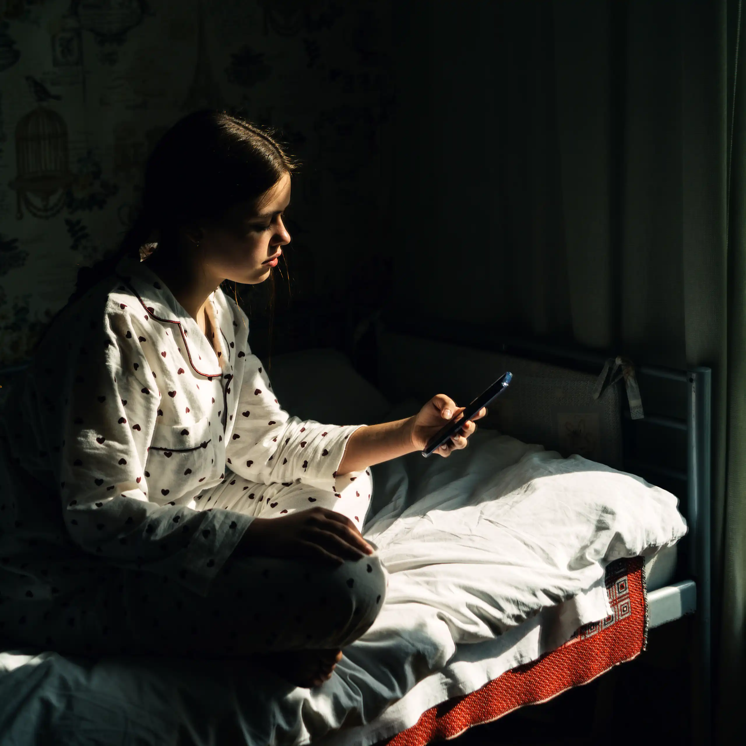 Uma adolescente vestindo pijama olha para o celular enquanto está sentada na cama em um quarto escuro