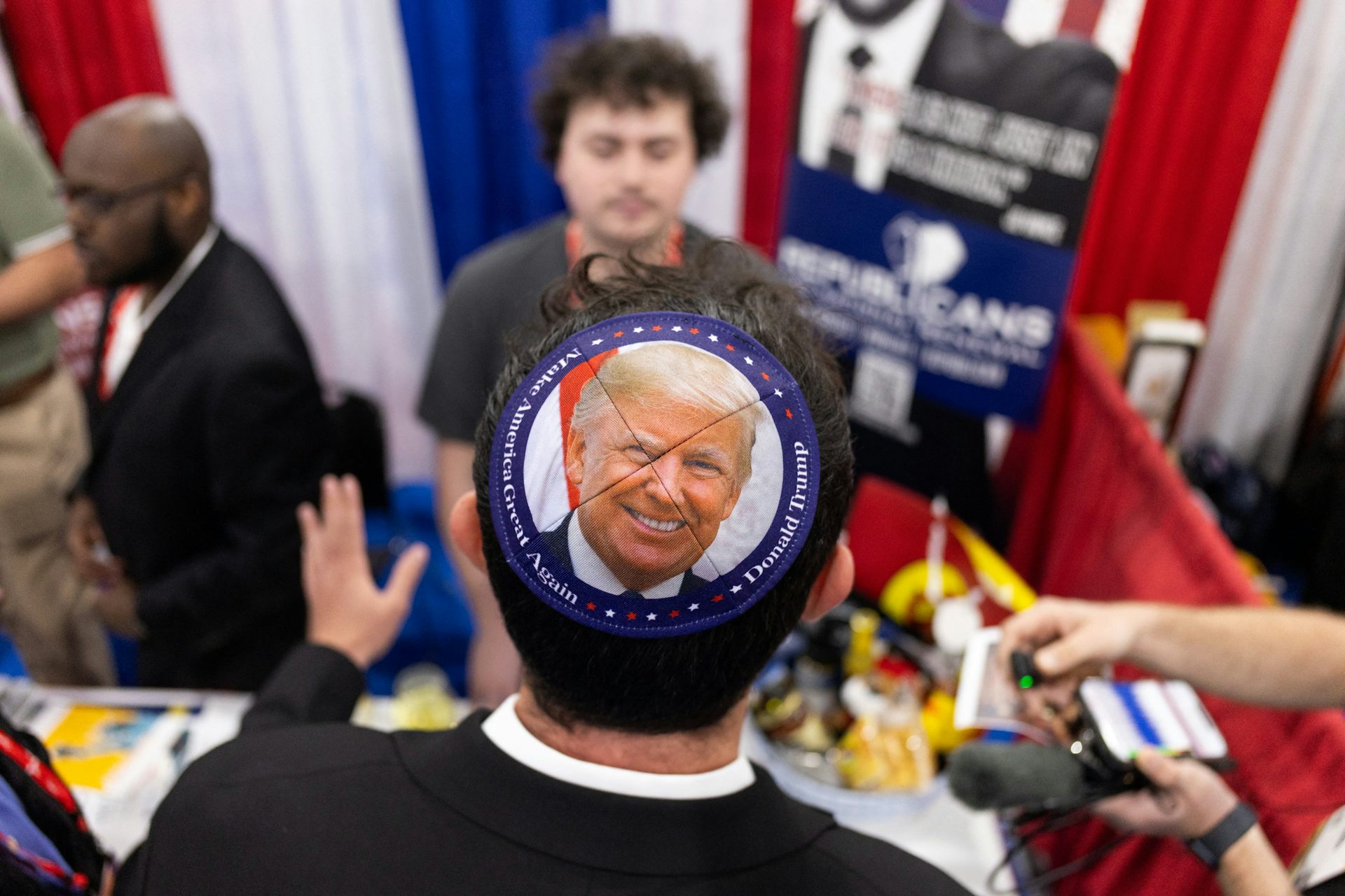 A man wearing a skullcap with a photo of Donald Trump on it stands at a table in a conference hall.