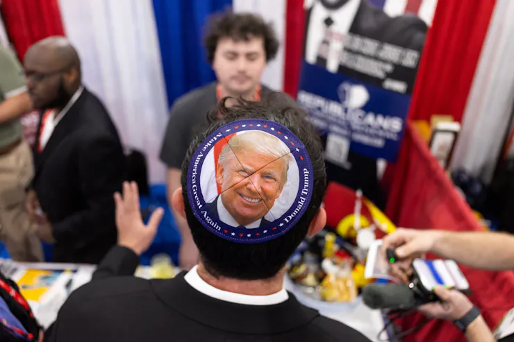 A man wearing a skullcap with a photo of Donald Trump on it stands at a table in a conference hall.