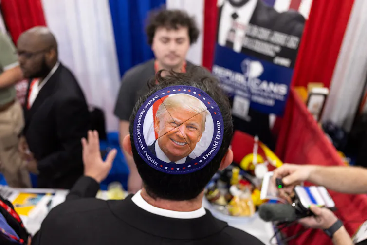 A man wearing a skullcap with a photo of Donald Trump on it stands at a table in a conference hall.