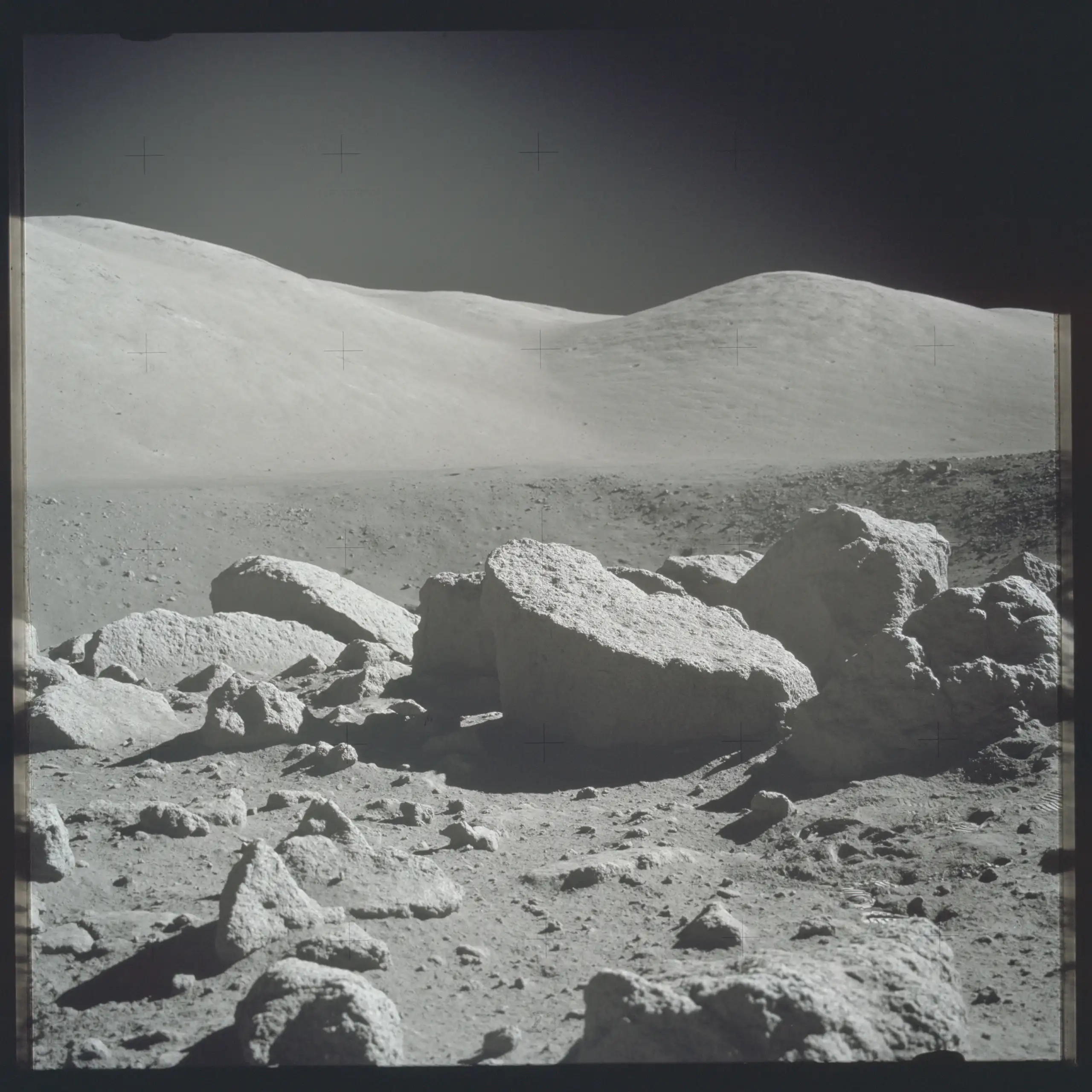 A black and white photo of the lunar surface, with several large rocks and a ridge in the background.