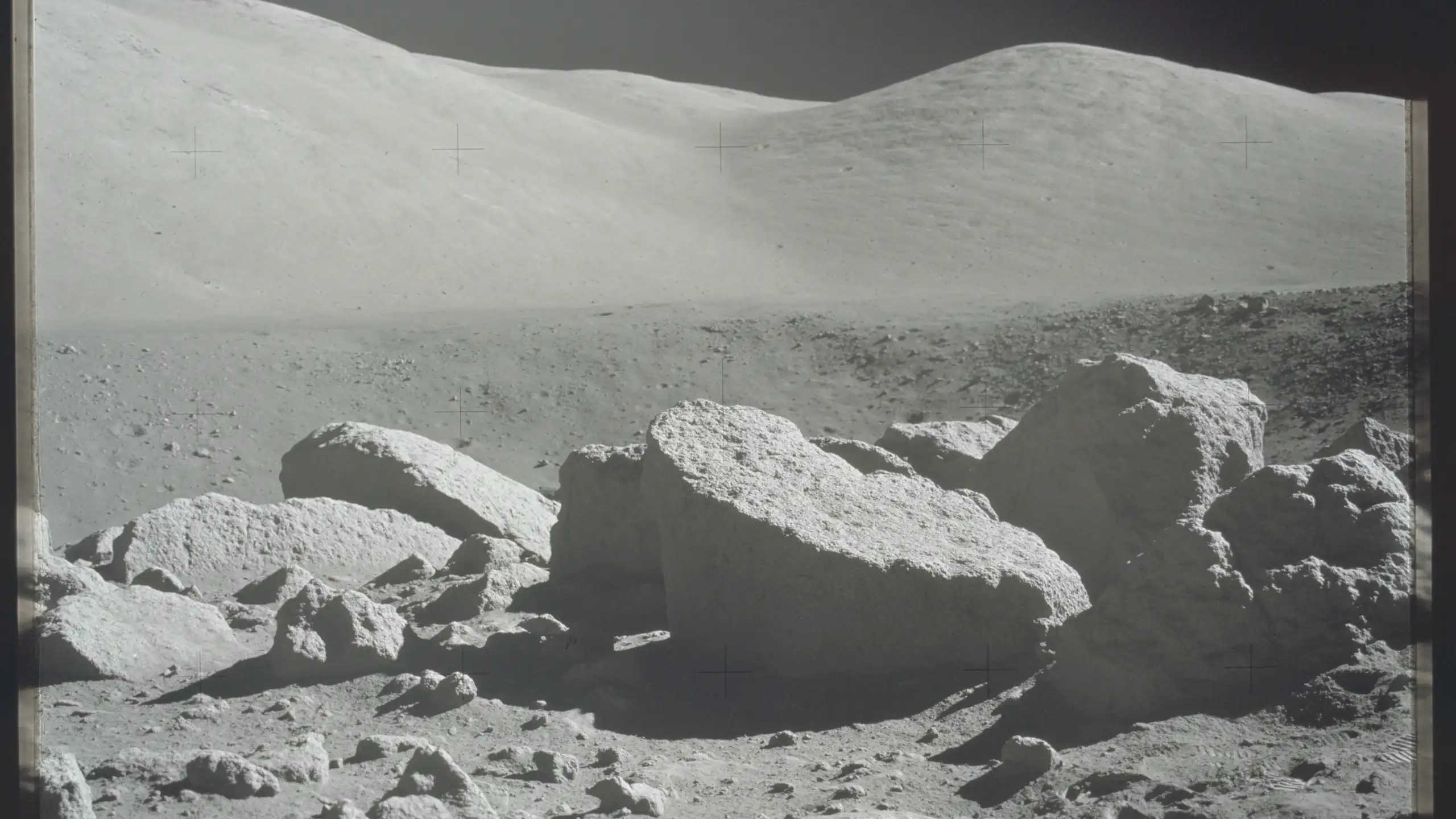 A black and white photo of the lunar surface, with several large rocks and a ridge in the background.