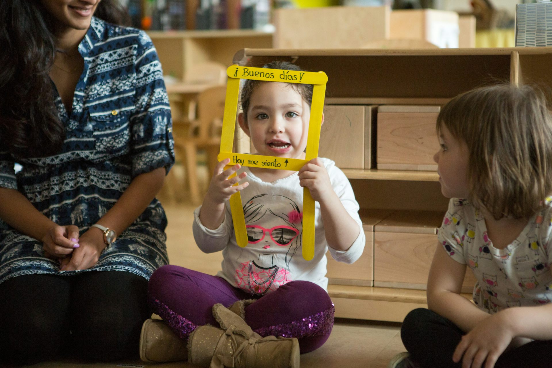 A seated child in a circle smiles and looks through a yellow square frame with the Spanish greeting buenas dias / hoy me sienta.
