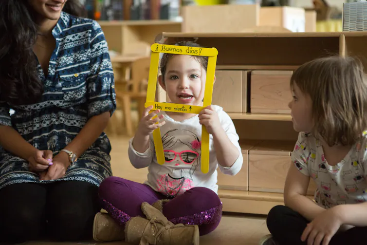 A seated child in a circle smiles and looks through a yellow square frame with the Spanish greeting buenas dias / hoy me sienta.