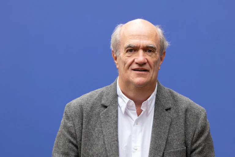 A middle-aged man with grey hair in a tweed jacket against a blue background.
