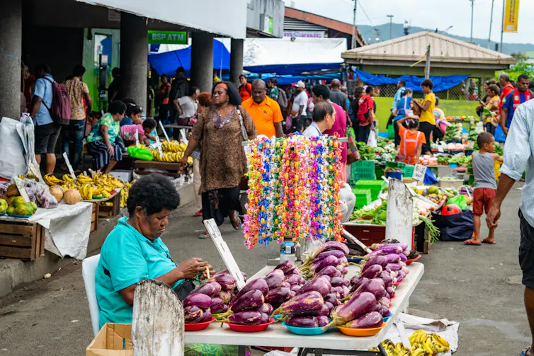 A street market with Fijian women staffing stalls selling fruit, floral garlands and vegetables