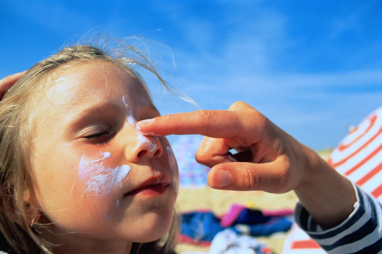 Adult applying sunscreen to girl's nose at beach, girl's eyes shut
