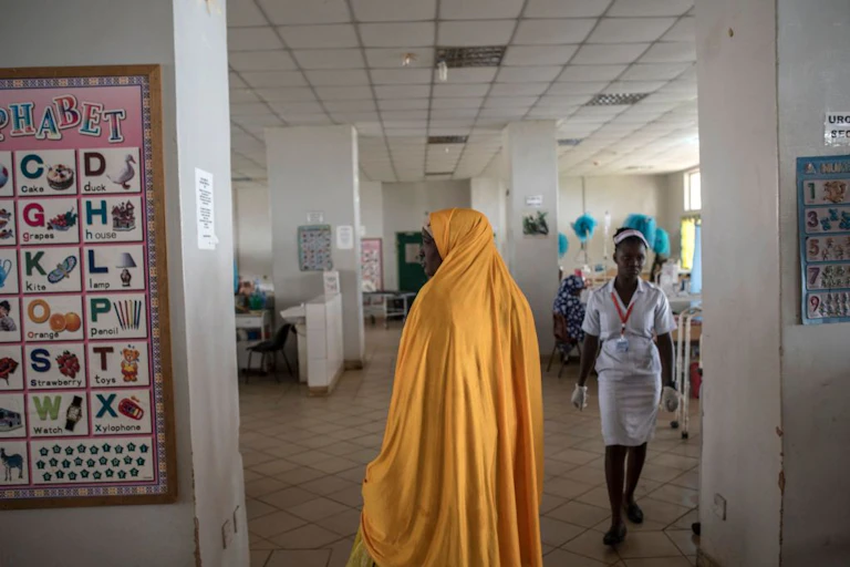 Woman in a robe stands in a hospital ward as a nurse approaches