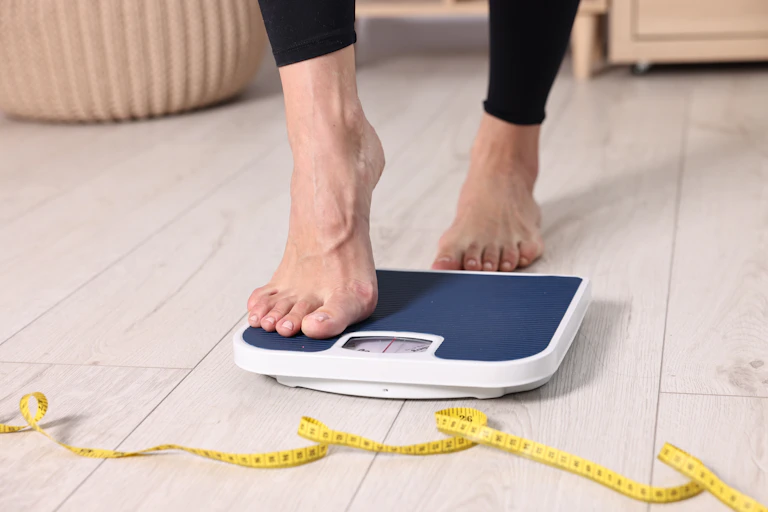 A picture of a person's feet, which are stepping onto a scale. There is a measuring tape on the floor in front of the scale.