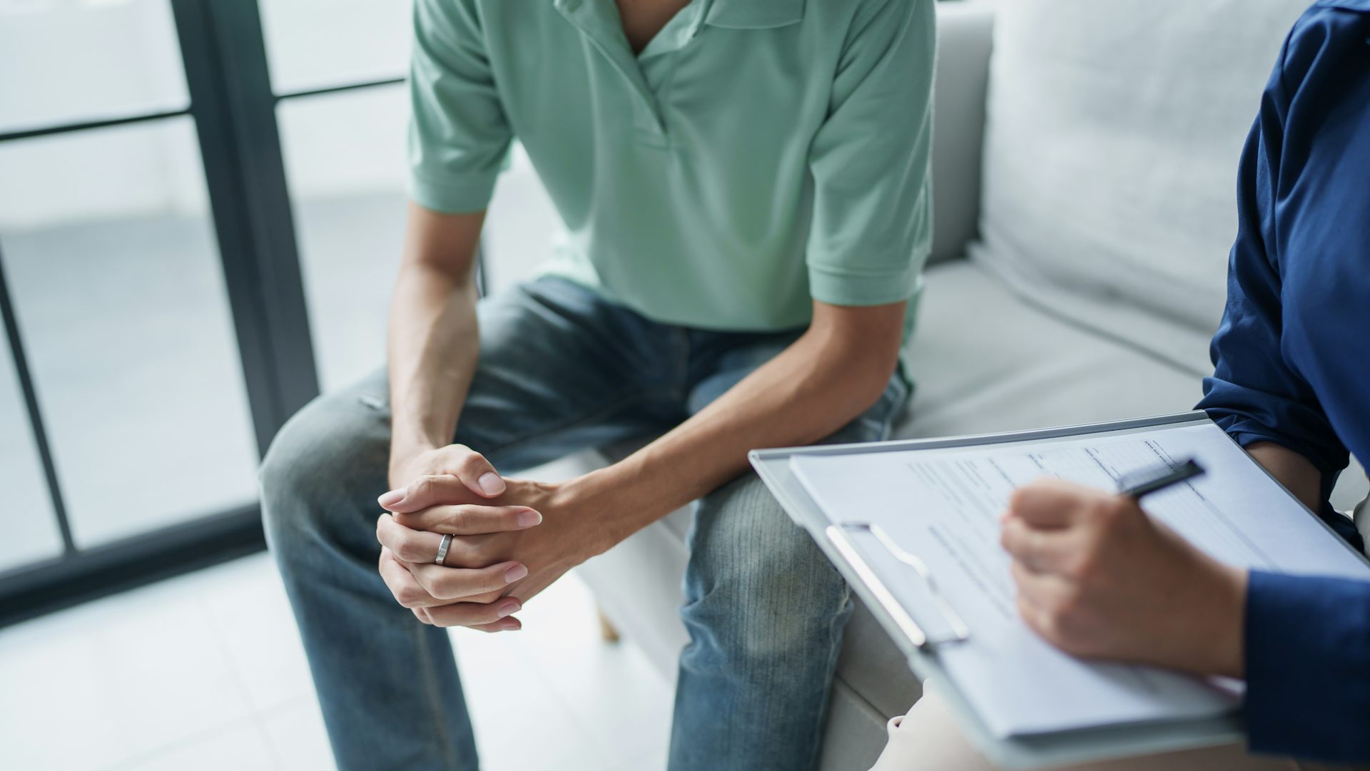 A man wearing a light teal shirt sits on a couch with his hands clasped. Sitting next to him is a person in a dark blue shirt who is writing on a clipboard with a pen.