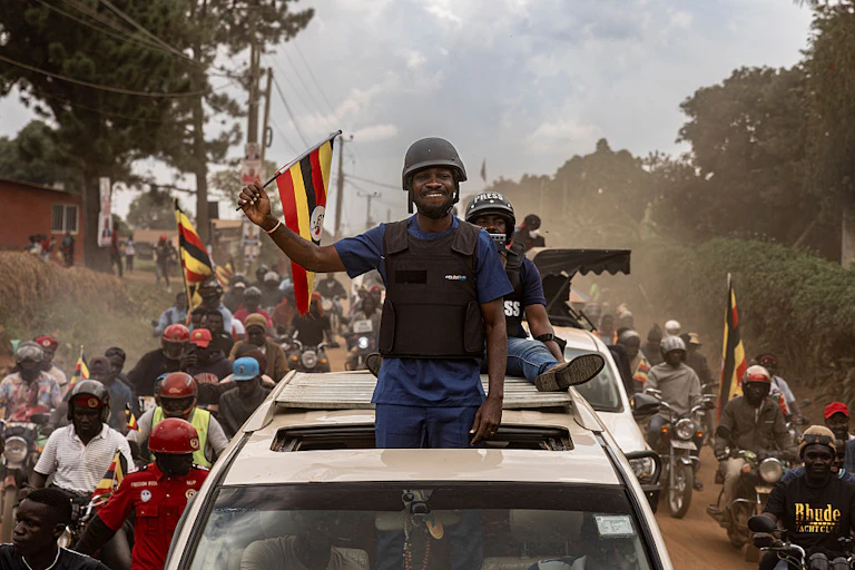 A man wearing a bulletproof vest standing from the sunroof of a vehicle and waving to a crowd of people, some of whom are waving flags.