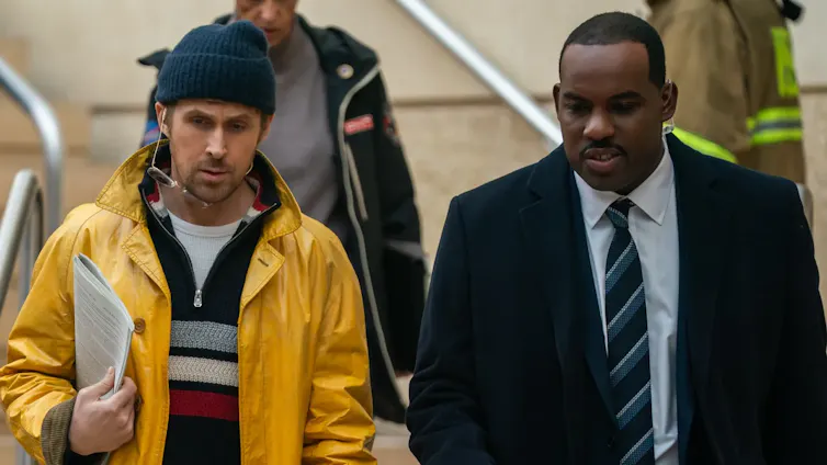 Ryland Grace, wearing a beanie and rain jacket, walks with Carl, wearing a suit jacket and tie.