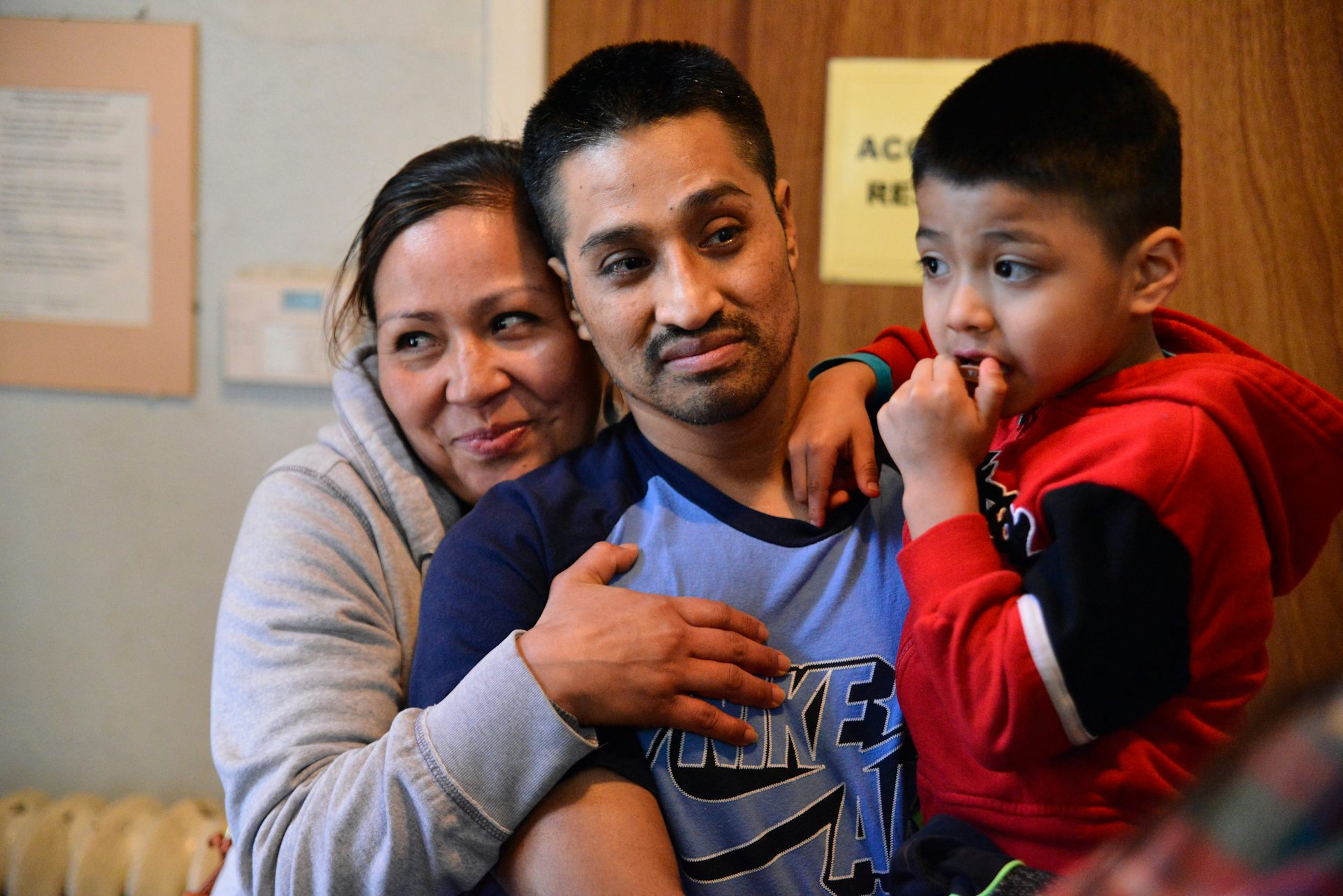 Man in blue shirt holds a child as woman hugs him from behind