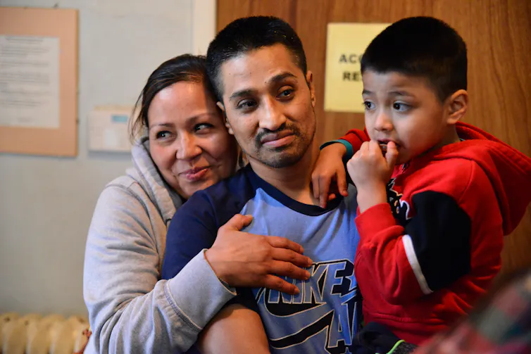 Man in blue shirt holds a child as woman hugs him from behind